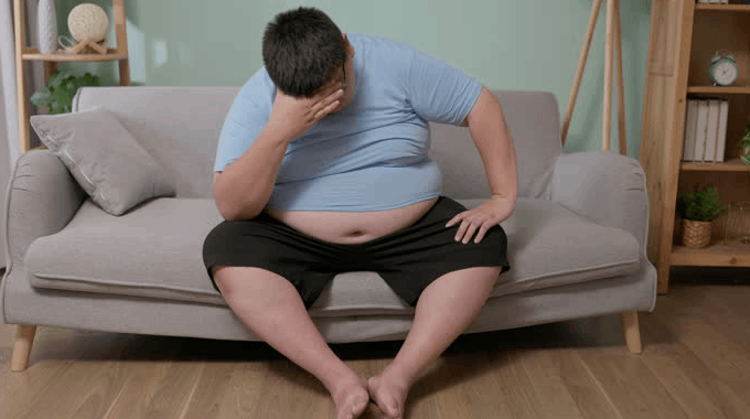 Adult man covering his face while sitting on a gray sofa with his shirt lifted over his abdomen.